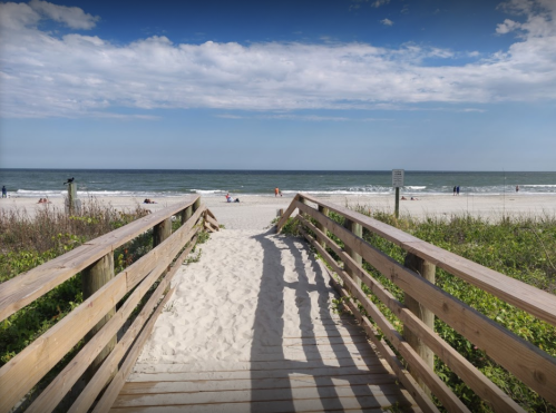 Wooden walkway leading to a sandy beach with gentle waves and people enjoying the sun under a blue sky.