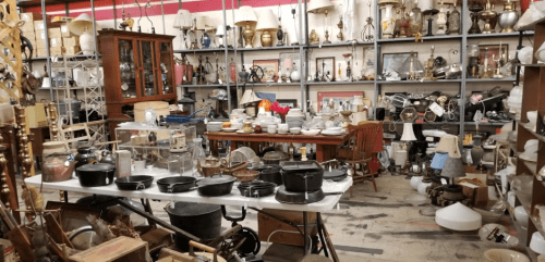 A cluttered antique shop filled with various items, including lamps, dishes, and furniture, on shelves and tables.