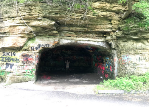A graffiti-covered rock cave entrance surrounded by greenery, with a person standing inside.