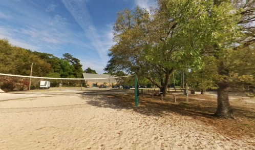 A sandy volleyball court surrounded by trees and a parking area under a clear blue sky.