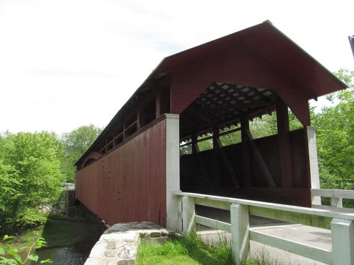 A red covered bridge spans a small stream, surrounded by green trees and a clear sky.