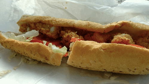A close-up of a sandwich filled with crispy fried chicken, lettuce, and tomato, served in a toasted hoagie roll.