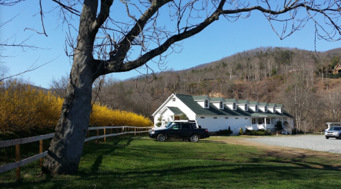 A white house with a green roof surrounded by trees and yellow bushes, set against a mountain backdrop on a clear day.