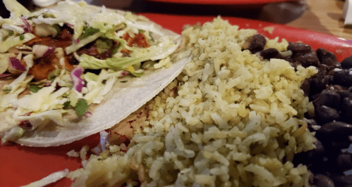 A plate featuring a fish taco with cabbage, cilantro, and a side of yellow rice and black beans.