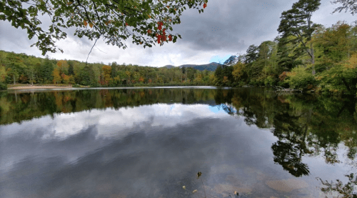 A serene lake surrounded by trees with autumn foliage, reflecting the cloudy sky and mountains in the background.