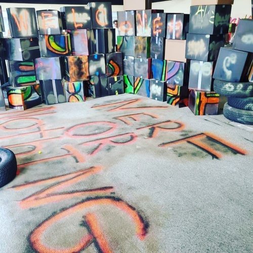 Colorful graffiti on cardboard boxes and a carpeted floor, with the word "WELCOME" prominently displayed.