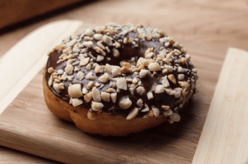 A chocolate-covered donut topped with crushed nuts, resting on a wooden surface.
