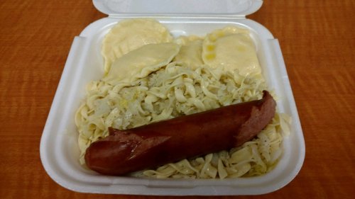 A takeout container with noodles, sauerkraut, and pierogi, topped with a sausage.