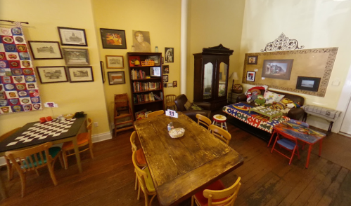 Cozy room with a wooden table, colorful chairs, bookshelves, and a sofa, decorated with artwork and vintage items.