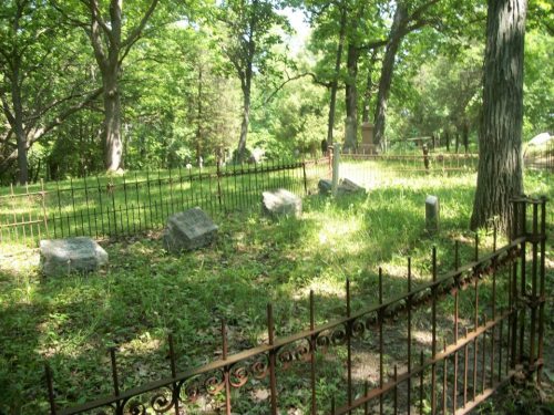 A serene cemetery with weathered gravestones surrounded by lush greenery and a wrought iron fence.