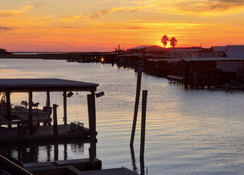Sunset over a calm waterway, with silhouettes of docks and palm trees against a vibrant orange and purple sky.