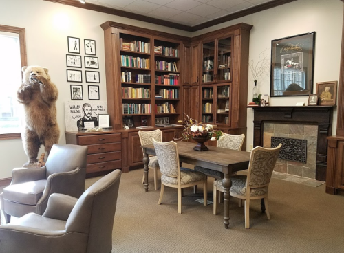 A cozy library with wooden shelves, a bear statue, a dining table, and comfortable chairs, featuring books and artwork.
