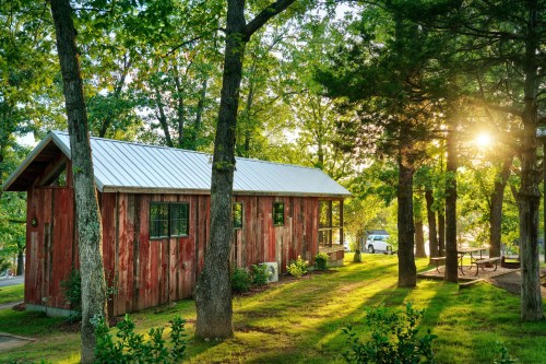 A rustic red cabin surrounded by trees, with sunlight filtering through the leaves and a picnic area nearby.