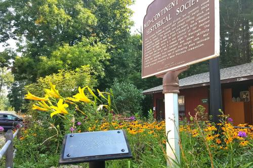 A historical marker stands near vibrant flowers in front of the Mid-Continent Historical Society building.