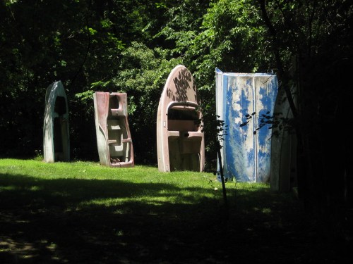 Five old, colorful boat hulls stand upright on green grass, surrounded by trees in a sunny outdoor setting.