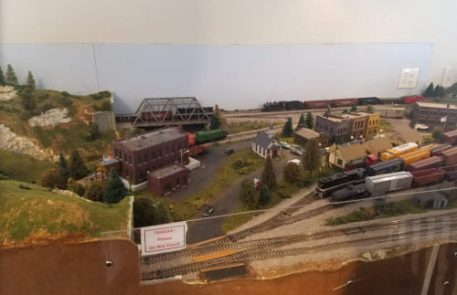A detailed model train layout featuring buildings, tracks, and a scenic landscape with trees and hills.