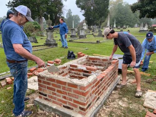 Two men work on a brick structure in a cemetery, with others in the background among gravestones.