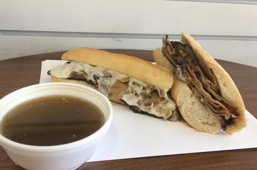 Two sandwiches filled with meat and cheese, served with a small cup of broth on a wooden table.