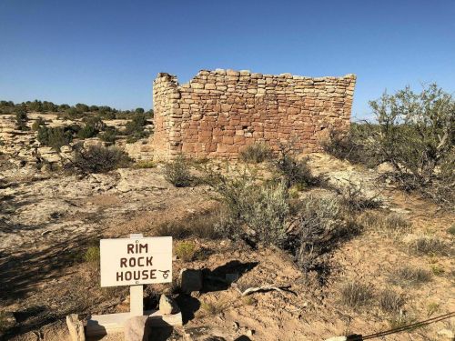 Ruins of a stone structure labeled "Rim Rock House" surrounded by desert vegetation under a clear blue sky.