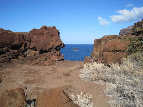 Rocky coastline with red cliffs framing a view of the blue ocean and distant island under a clear blue sky.