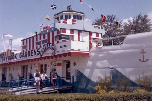 A colorful building resembling a ship, decorated with flags, with people standing outside in a garden setting.