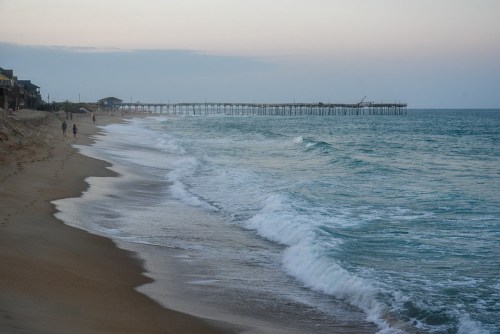 A serene beach scene with gentle waves, a wooden pier in the distance, and a few people walking along the shore.