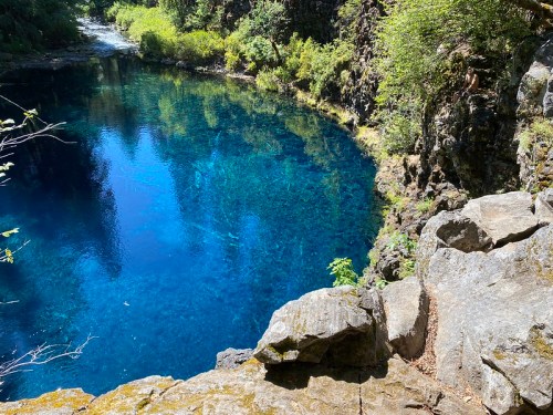 A serene blue pond surrounded by lush greenery and rocky cliffs, reflecting the clear sky above.