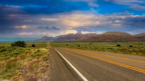 A scenic road stretches through a green landscape, with mountains and stormy skies in the background. Lightning is visible.