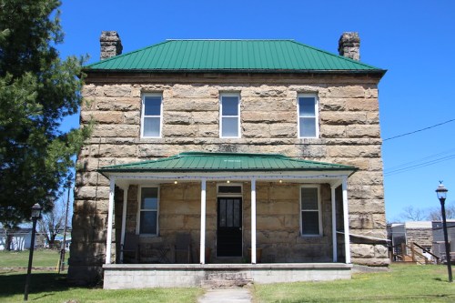 Historic stone house with a green metal roof, front porch, and clear blue sky in the background.