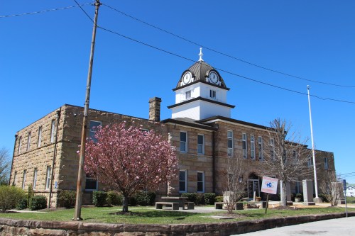 Historic stone building with a clock tower, surrounded by trees and a clear blue sky.