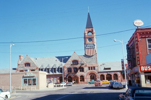 Historic train station with a clock tower, featuring red stone architecture and nearby vintage cars and buildings.