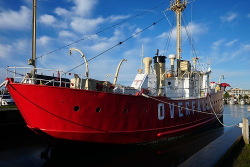 A red lighthouse ship named "Overfall" docked in a calm harbor under a blue sky with scattered clouds.