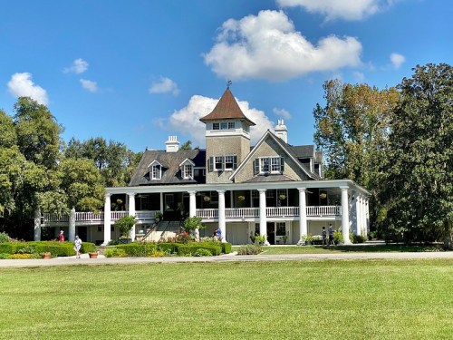 A large, elegant house with a tower, surrounded by greenery and blue skies, featuring a spacious front porch.