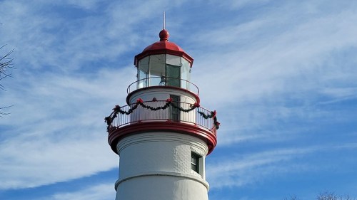 A white lighthouse with a red top, decorated with garlands, against a blue sky with wispy clouds.