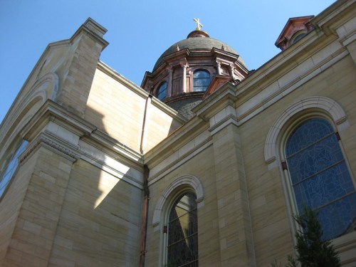A low-angle view of a building with a dome and cross, featuring stone walls and stained glass windows against a blue sky.