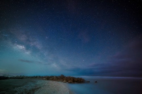 A serene beach at night, with a starry sky and the Milky Way visible above calm ocean waves and rocky formations.