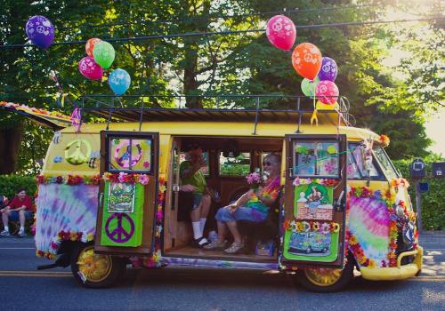 A colorful, decorated yellow van with flowers and balloons, parked on a sunny street with people inside.