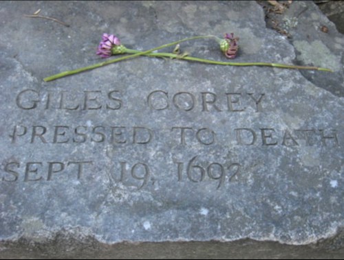A gravestone engraved with "Giles Corey, pressed to death, Sept 19, 1692," adorned with a flower.