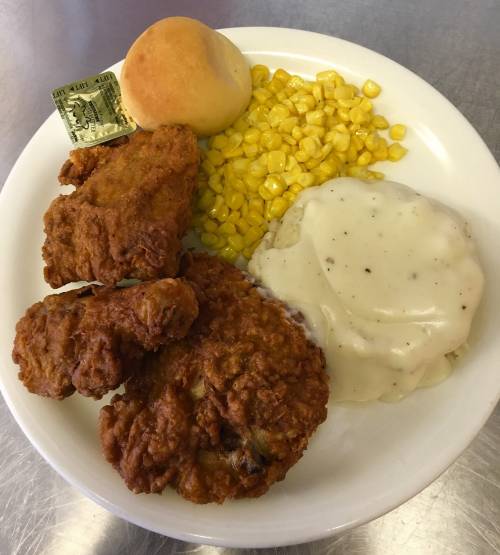 A plate of fried chicken, corn, mashed potatoes with gravy, a roll, and a dollar bill.