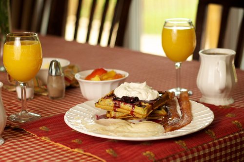 A breakfast table featuring waffles with berries, bacon, eggs, fruit, and mimosas on a red checkered tablecloth.