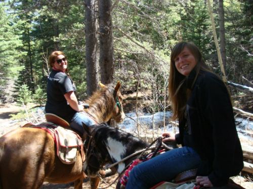 Two women on horseback in a forested area, smiling and enjoying a sunny day outdoors.