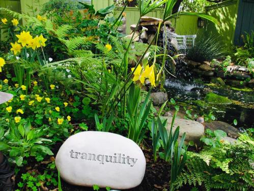 A serene garden scene featuring yellow flowers, lush greenery, and a stone marked "tranquility" near a small waterfall.