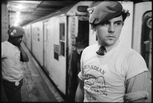 Two individuals in berets and t-shirts stand in a subway station, one looking away while the other is in the background.