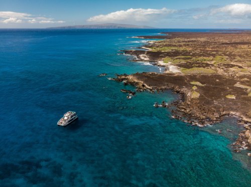 Aerial view of a boat near a rocky coastline with clear blue waters and distant land under a partly cloudy sky.