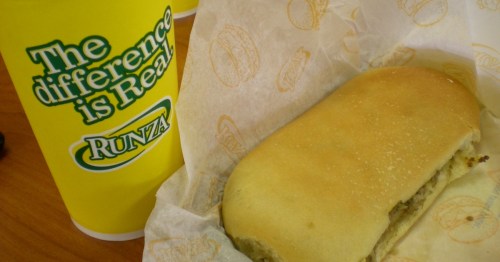 A Runza sandwich on a paper wrapper next to a large yellow cup with the text "The difference is Real."
