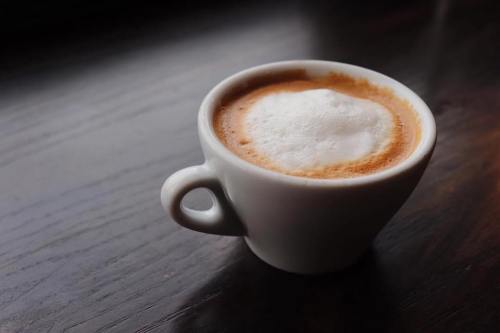 A small white cup filled with espresso topped with frothy milk, resting on a dark wooden surface.