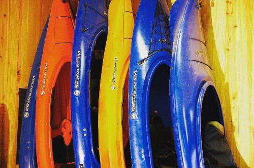 Four colorful kayaks (orange, yellow, blue) are neatly arranged against a wooden wall.