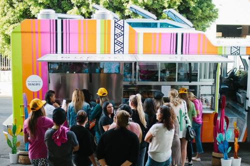 A colorful food truck with a line of people waiting to order, surrounded by vibrant decorations and cacti.