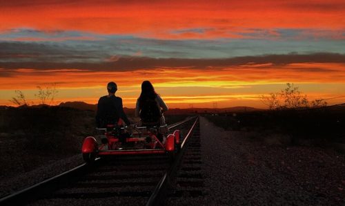 Two people on a pedal cart on railway tracks, silhouetted against a vibrant sunset sky.