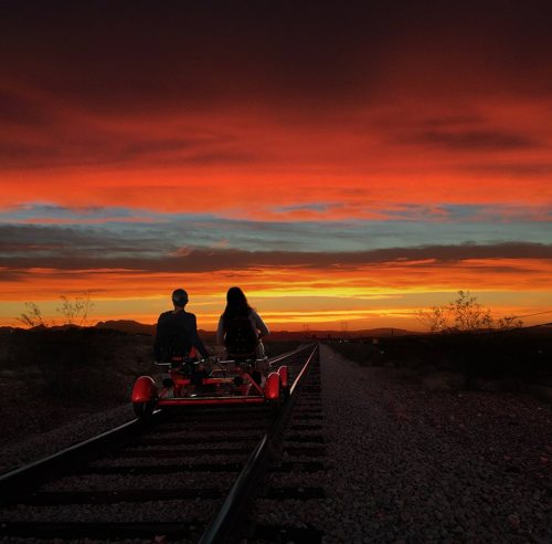 Two people ride a red pedal cart on railway tracks, silhouetted against a vibrant sunset sky.